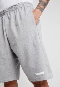 Shorts en coton gris avec une ceinture élastique, arborant un logo blanc "hummel" sur la jambe gauche. Le tissu a une texture lisse.