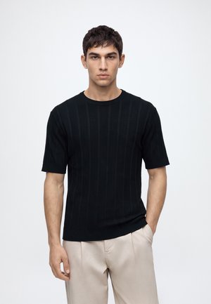 Jeune homme aux cheveux courts et foncés, portant une chemise noire à manches courtes et un pantalon beige clair, debout devant un fond blanc uni.
