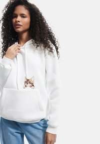 Sudadera con capucha blanca, fabricada en un tejido suave, con un diseño de gato bordado en detalle en el bolsillo frontal. Incluye un bolsillo tipo canguro.