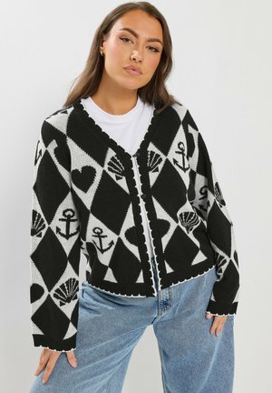 Vrouw draagt een zwart-wit geruit vest met knopen, met ankers, schelpen en harten, over een wit T-shirt en blauwe spijkerbroek.