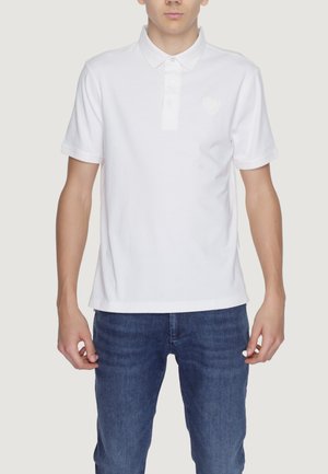 Polo shirt bianco in cotone con colletto classico, patta con tre bottoni e un logo discreto sul lato sinistro del petto. Abbinato a jeans blu scuro.