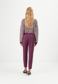 Woman with long blonde hair wearing a striped maroon and white sweater, purple pants, and black heeled shoes, standing against a white background.
