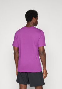 Lilla kortærmet t-shirt med rund hals, lavet af blødt stof. Båret med sorte shorts, vist fra ryggen, der afslører det enkle design.