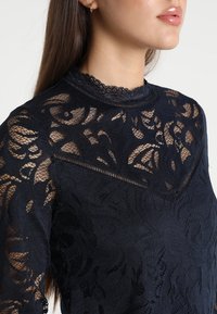 Marinblus i spets med hög hals och scalloped detaljer, intrikata blommönster och genomskinliga partier längs ärmarna och bröstet.