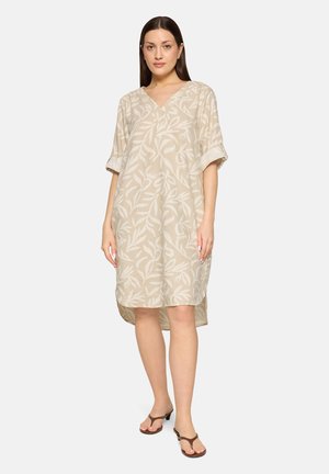 Frau trägt ein beige knielanges Kleid mit weißem Blattmuster, V-Ausschnitt, hochgekrempelten Ärmeln und braunen Sandalen mit Absatz, steht vor weißem Hintergrund.
