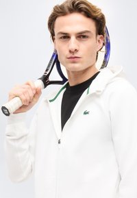 Νεαρός άνδρας που φοράει λευκή φούτερ με φερμουάρ, κρατώντας ρακέτα τένις που ακουμπά στον ώμο του, κοιτάζοντας μπροστά με ουδέτερη έκφραση.