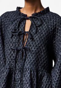 Robe bleu marine texturée avec un col à volants, un lien à nouer au niveau du cou, et des manches bouffantes. Présente une taille cintrée et un tissu à motif subtil.