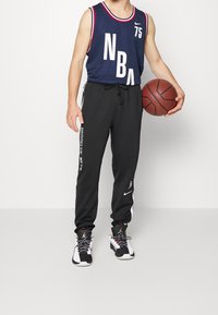 Sininen NBA-hihaton paita valkoisilla grafiikoilla ja punaisilla yksityiskohdilla, yhdistetty mustiin joggereihin, joissa on valkoinen "BROOKLYN NETS" -teksti ja sivuraidat.