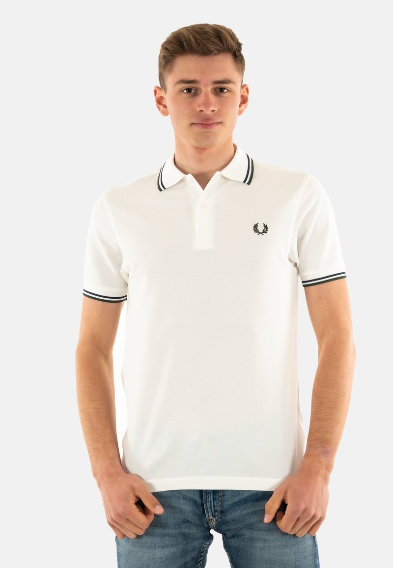 Polo bianco con colletto e polsini a righe nere e verdi, caratterizzato da un piccolo emblema nero sul petto sinistro. Indossato con jeans blu.
