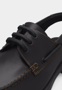 Μαύρο δερμάτινο παπούτσι τύπου bateau με καφέ ραφές και χοντρές θηλιές κορδονιών, που δείχνει λεπτομερή κοντινή εικόνα του μπροστινού και πλευρικού τμήματος.