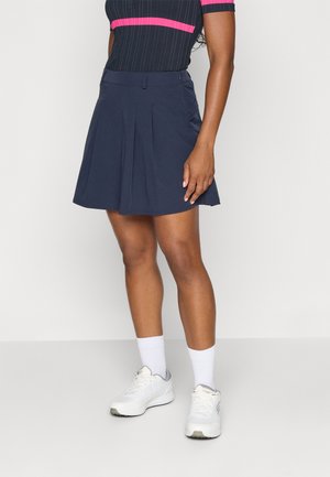 Falda plisada azul marino hecha de tejido ligero, con una cintura ajustada y un ligera abertura. Combinada con zapatillas blancas y calcetas hasta el tobillo.