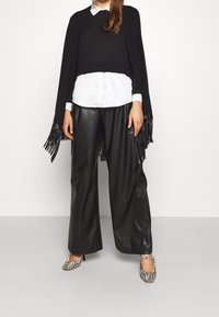 Donna che indossa un cappotto nero con frange, una camicia bianca con bottoni, pantaloni in pelle neri larghi e scarpe con tacco a stampa zebra, in piedi su un pavimento grigio.
