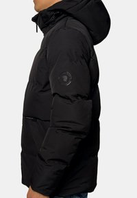 Veste doudoune noire avec capuche, en tissu texturé, et un logo rond sur la manche gauche ; elle possède une fermeture éclair sur le devant et des poches latérales.