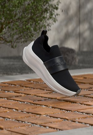 Μαύρο sneaker slip-on με λευκή σόλα και λογότυπο Calvin Klein στο λουράκι, τοποθετημένο πάνω από ξύλινα πλακίδια εξωτερικού χώρου.
