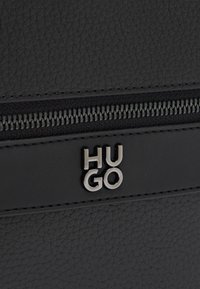 Μαύρη τσάντα από δερμάτινη υφή με κορυφαίο φερμουάρ, μαύρα μεταλλικά στοιχεία και μεταλλικό λογότυπο με τα γράμματα "HUGO" τοποθετημένο στο κέντρο πάνω σε λείο δερμάτινο λουρί.