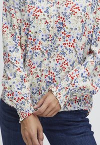 Blusa a maniche lunghe floreale color crema con motivi rossi, blu e verdi. Presenta chiusura con bottoni e polsini arricciati per un tocco di dettaglio.