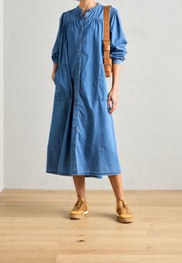 Denim skjortekjole med lange puffærmer, knaplukning fortil, sidelommer og en løs pasform. Bæres med brune sneakers med striber.