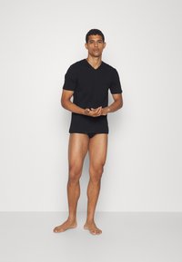 T-shirt noir à col en V à manches courtes en tissu doux, associé à un slip noir assorti. Le modèle se tient pieds nus contre un fond uni.