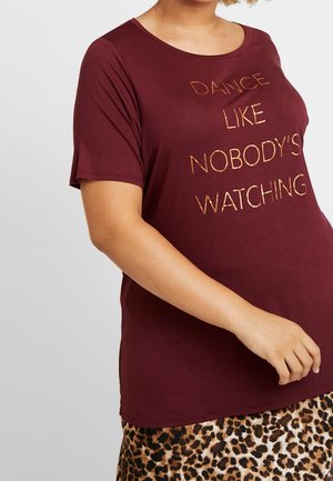 Osoba nosząca bordową koszulę z napisem „Dance Like Nobody's Watching” i spódnicę w panterkę, na gładkim jasnoszarym tle.