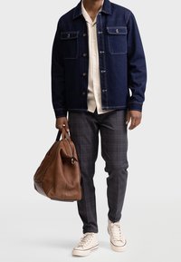 Denim jacket with dark blue color, two chest pockets, beige shirt, plaid trousers, and brown leather duffle bag. Tan sneakers complete the look.