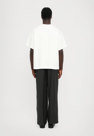 Λευκή κοντομάνικη μπλούζα με ίσια στρώση και λεπτομέρειες ραφής, συνδυασμένη με φαρδιά σκούρα γκρι παντελόνια, θέα από πίσω.