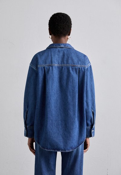 Denim shirt in dark blue, relaxed fit with a pointed collar, dropped shoulders, and buttoned cuffs. Back view shows curved hemline and stitching.