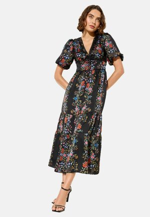 Kobieta w czarnej sukience midi z kolorowym kwiatowym nadrukiem, bufkami na rękawach, dekoltem w serek i czarnych sandałach na wysokim obcasie z paskiem wokół kostki, stoi z rękami za plecami.