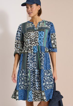 Femme portant une robe patchwork mi-longue aux genoux avec des motifs bleus, noirs, blancs et une casquette bleu marine, tenant un sac bleu marine dans sa main gauche.