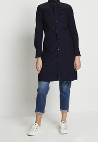 Person som bär en långärmad, knäppt marinblå skjortklänning över uppvikta blå jeans och vita sneakers, stående mot en plain bakgrund.