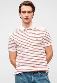 Polo à manches courtes blanc avec des rayures horizontales rouges. Il comprend un col classique, une patte à deux boutons et un logo sur le côté gauche de la poitrine.