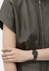 Ronde zwarte polshorloge met een gestructureerde band, gecombineerd met een groene leren kledingstuk met een ritssluiting en een gladde oppervlakte.