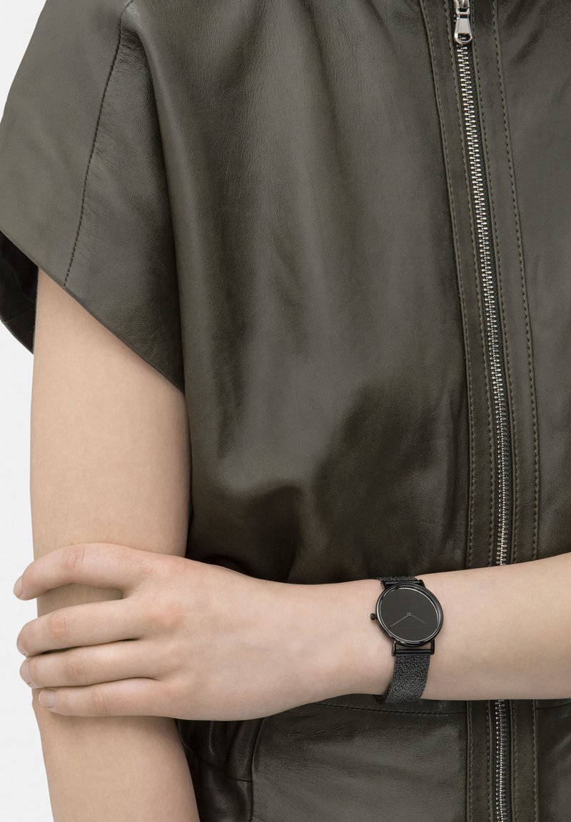 Ronde zwarte polshorloge met een gestructureerde band, gecombineerd met een groene leren kledingstuk met een ritssluiting en een gladde oppervlakte.