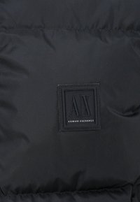 Matière de doudoune noire avec texture matelassée et patch logo carré brodé affichant "AX" et "ARMANI EXCHANGE" en texte blanc.