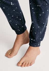 Marineblauwe pyjamabroek met witte sterprint, met elastische boorden bij de enkels. Naakte voeten zijn zichtbaar, met een natuurlijke huidstructuur.