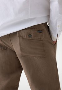 Ruske hlače od čvrstog materijala s stražnjim džepom i crnim oznakom "GUESS". Imaju detalj gumba blizu džepa.