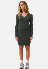 Abito lavorato a maglia verde scuro con scollatura a V, vestibilità slim, maniche lunghe e polsini a coste. Indossato con sneakers grigie e calze bianche.