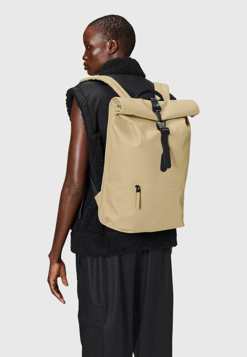 Zaino beige a roll-top con una tracolla nera. Presenta una tasca frontale con zip e una finitura matte liscia. Progettato con spallacci imbottiti.