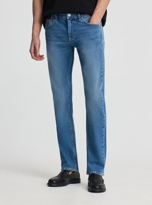 Uomo che indossa jeans blu a gamba dritta e mocassini neri, con una mano in tasca e l'altra rilassata lungo il fianco.