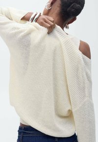 Helle, cremefarbene gestrickte Pullover mit einem offenen Rücken-Design, gerippten Bündchen und einem lockeren Schnitt, kombiniert mit einem dunklen Taillenbund. Silberne Creolen.
