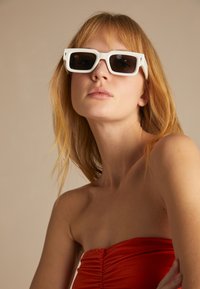 Gafas de sol rectangulares blancas con lentes oscuros, que cuentan con un diseño de marco grueso y bordes definidos. El modelo tiene el cabello largo y rubio y lleva una camiseta roja.