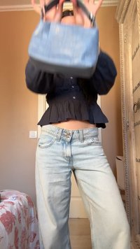 Persona che indossa jeans azzurri chiari e top nero, tiene sollevata una piccola borsa azzurra, in piedi all'interno vicino al letto e a una porta in legno decorativa.