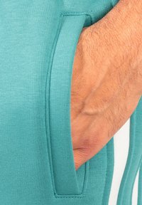 T-shirt de sport teal avec une poche à texture et des coutures visibles, montrant un gros plan du tissu et de la main.