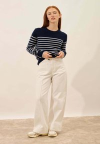 Sweter w marynarskie paski z białymi paskami, wysoki kołnierz; szerokie kremowe spodnie z przednimi kieszeniami, kontrastowe przeszycia; jasne buty.