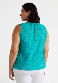 Una donna in un top senza maniche color teal con dettagli in pizzo e un collo arricciato, abbinato a dei pantaloni bianchi.