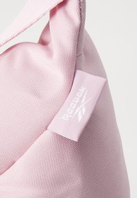 Sac en tissu texturé rose avec une forme arrondie, présentant une étiquette du logo Reebok en blanc. Des détails de couture visibles embellissent son design.