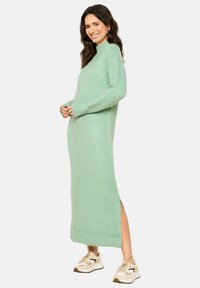 Mentolno zelena, dolga obleka z dolgimi rokavi, pletena v rebrasti vzorec, z visokim ovratnikom in bočnimi razporki. Oblečena z nevtralno obarvanimi športnimi copati, mehka tekstura.