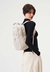 Beige Rucksack mit glatter Stoffstruktur, ausgestattet mit gepolsterten Schultergurten, einer Fronttasche und dekorativen Hardware-Akzenten.