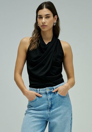 Femme aux longs cheveux bruns portant un haut sans manches noir drapé et un jean taille haute bleu clair, debout avec les mains dans les poches.