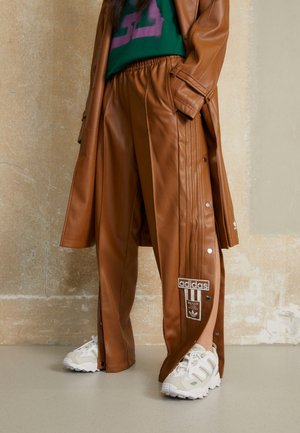 Osoba ubrana w brązowe skórzane spodnie dresowe Adidas z zatrzaskami po bokach, białe trampki i dopasowany brązowy skórzany płaszcz na zielono-fioletowej bluzce.