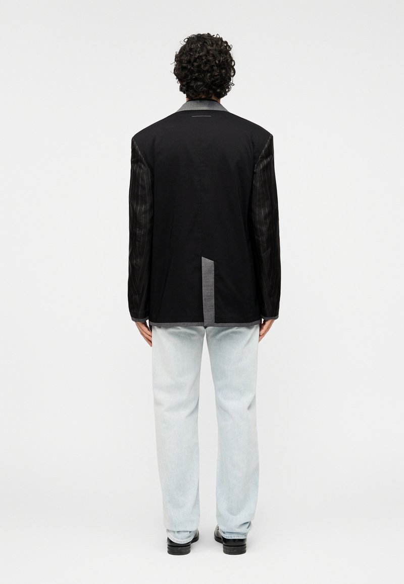 Veste noire avec manches en tissu gris à motifs, col à revers cranté et une fente à l'arrière. Jeans délavés clairs complètent la tenue.